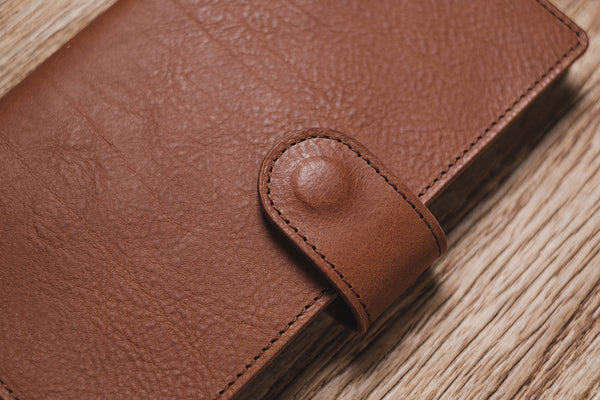 A brown pebbled leather traveler's notebook with a strap closure, displayed closed, close up of snap closure and stitching
