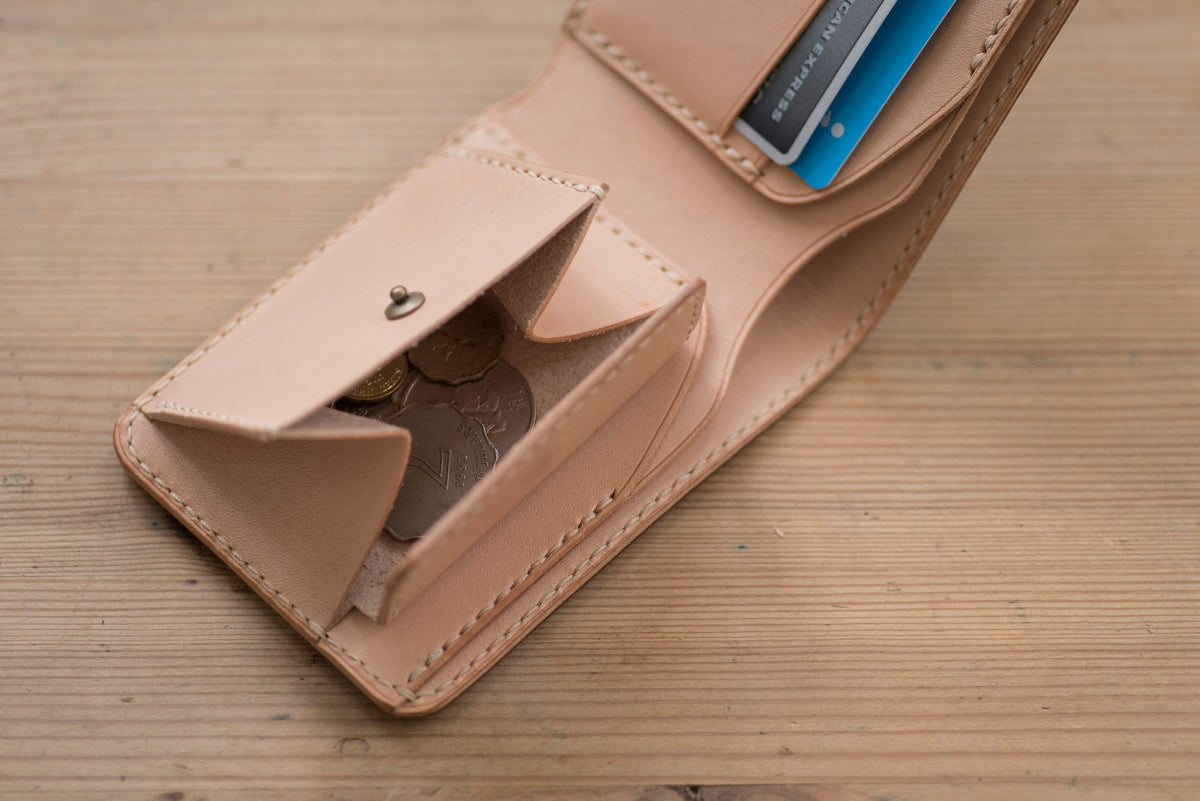 A natural leather wallet with a flap open, revealing a small coin pocket with coins inside and 2 card slots with 2 cards.