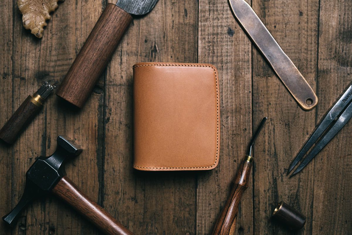 A caramel leather bifold wallet with visible stitching along the edges. The wallet is surrounded by various metal and wooden hand tools, including a hammer, a creaser, a knife, and a pair of dividers, all arranged on a wooden surface.
