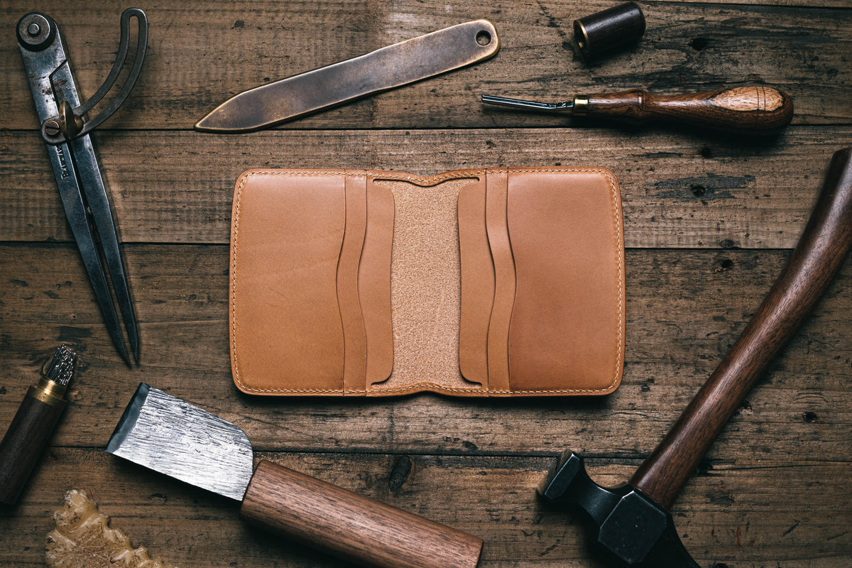 A caramel Buttero leather billfold wallet laid out flat on a wooden surface, surrounded by tools such as a knife and hammer, indicative of its handcrafted making.