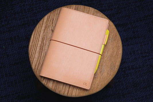 A natural leather bound journal with a yellow pen tucked into the elastic band. The journal has the initials "CL" embossed on the front cover.