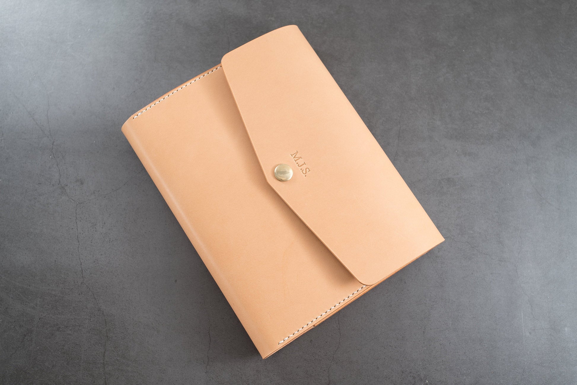 A natural trifold leather notebook cover with a flap closure and a gold button. The notebook cover has the initials "M.J.S." embossed on the front.