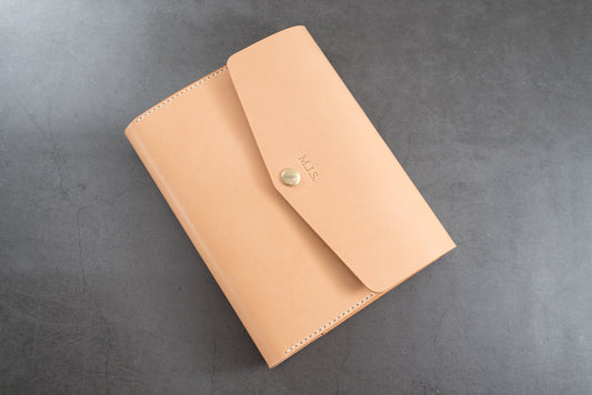 A natural trifold leather notebook cover with a flap closure and a gold button. The notebook cover has the initials "M.J.S." embossed on the front.