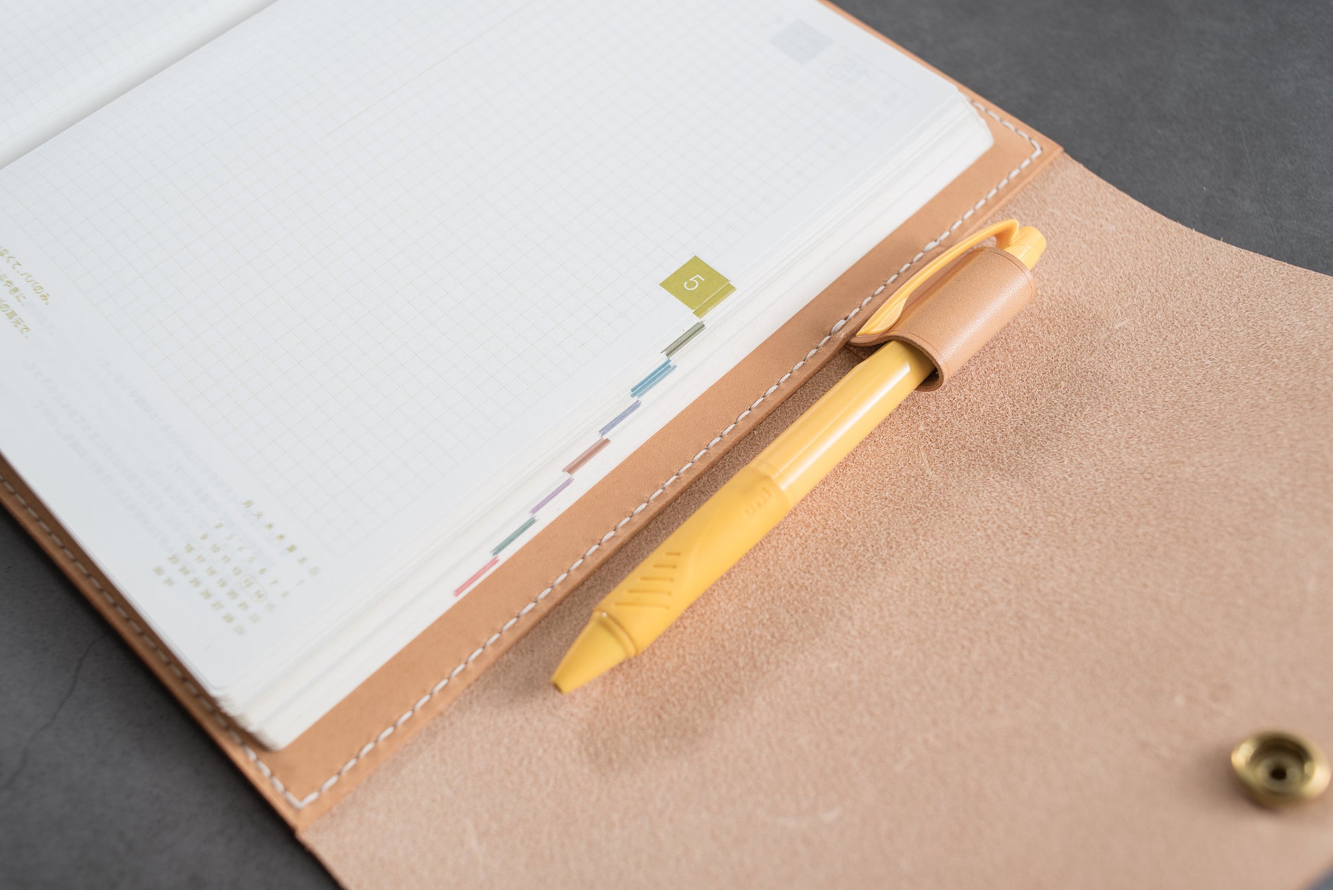 A natural leather notebook cover with a yellow pen tucked into the cover. The notebook has a grid pattern on the pages and a small number "5" on the side.