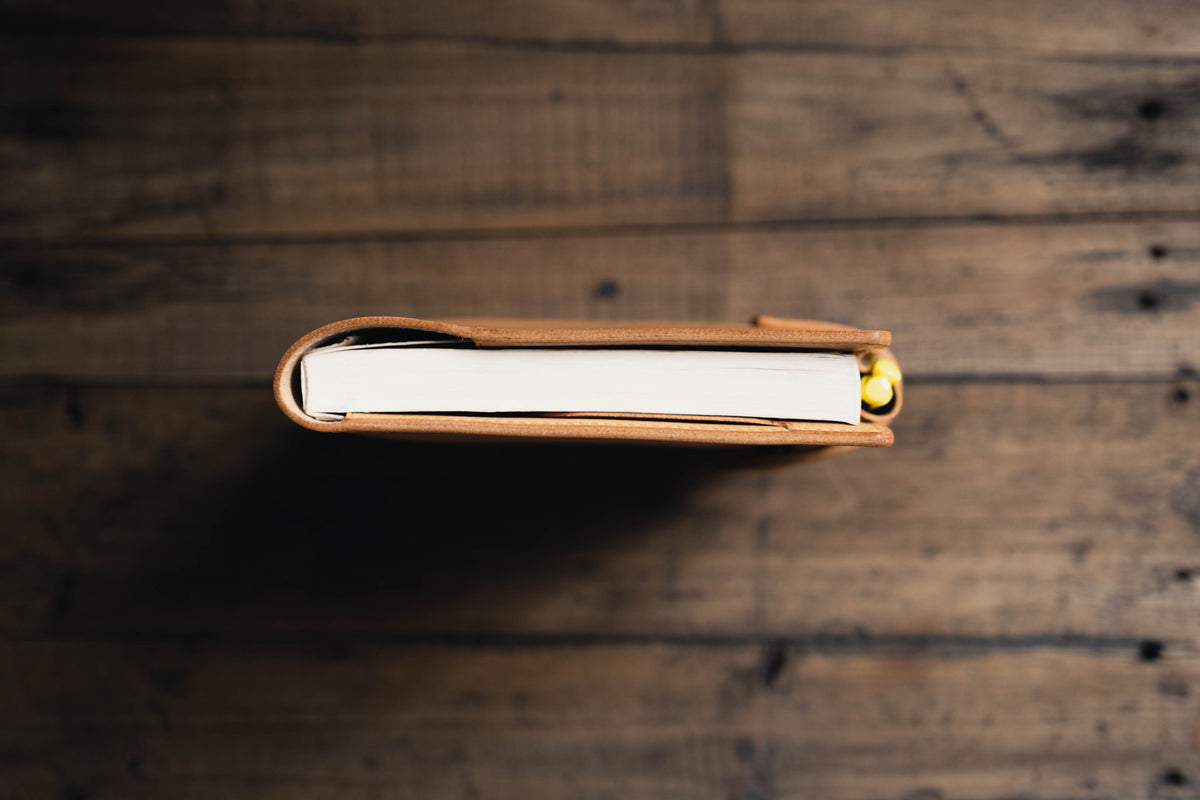 A natural leather notebook cover with a smooth finish, featuring a wrap-around design that secures a white paper notebook. A small yellow pen or pencil is tucked into the side of the cover, visible against the white pages of the book.