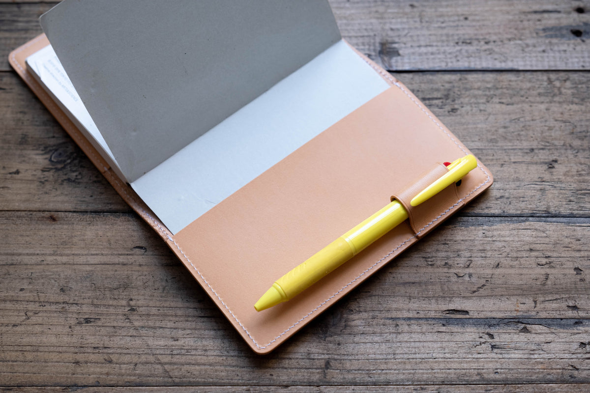 A natural leather notebook cover with white stitching, featuring an integrated pen loop holding a bright yellow multi-color ballpoint pen. The cover is open, revealing a plain white paper notebook insert tucked inside the leather sleeve.