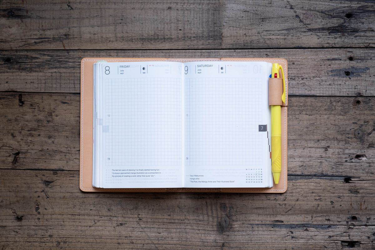 An open planner with a natural leather cover featuring a grid-lined layout for Friday and Saturday. A bright yellow multi-color ink pen is held in a matching leather pen loop attached to the side of the planner cover.