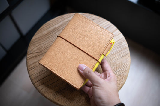 A natural leather notebook cover with a textured surface and a brown elastic band closure. A yellow pen is visible inside the journal.