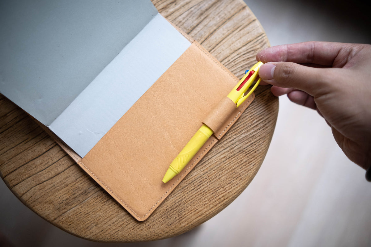 A natural leather notebook cover with a yellow multi-color pen in a pen holder. The notebook is open, revealing gray and white pages. The pen has red, blue, and black ink options. 