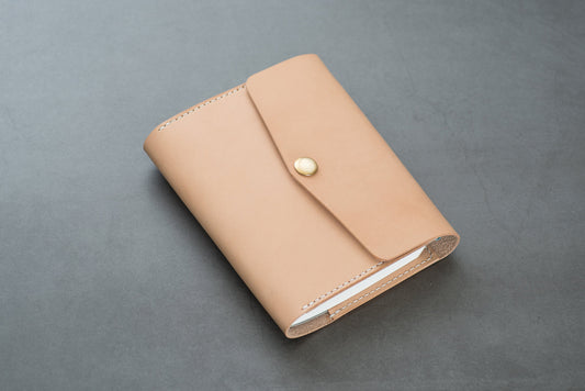 A natural A6 trifold leather notebook cover with a natural leather texture, a pen loop, and a front closure strap.
