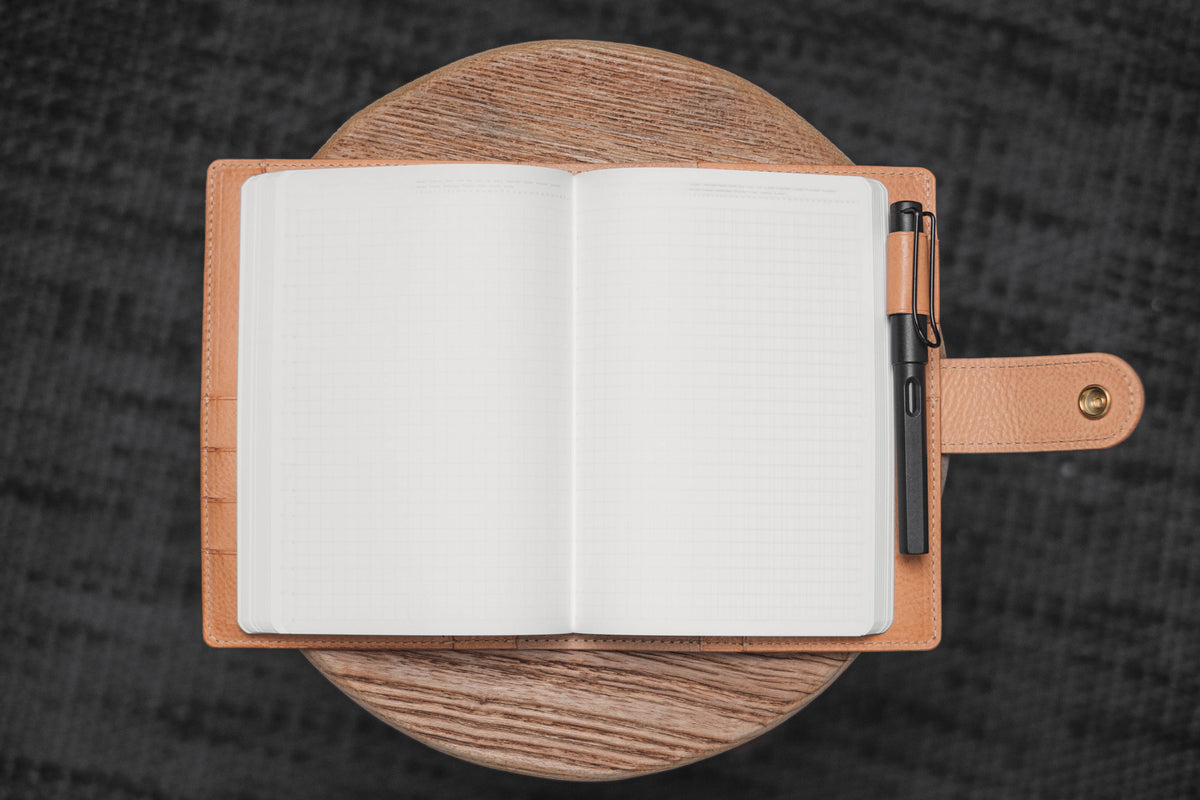 A natural leather journal cover with a black pen tucked into the cover. The journal is open to a blank page with lined paper. 