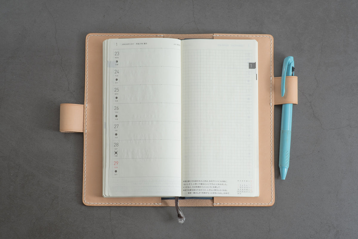 A natural leather notebook cover with an interlocking closure and a stitched edge. The notebook is open to reveal a white page with a dotted pattern.