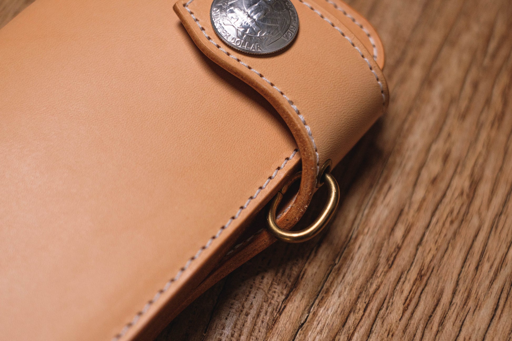 A close-up of a natural leather wallet with a silver coin attached to the front. The wallet has a brass ring attached to the side.