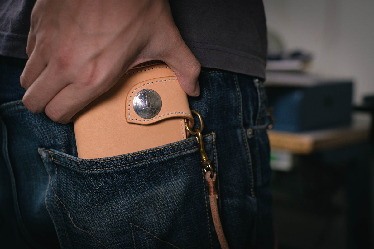 A natural wallet with a silver metal button and a brown leather strap attached to a metal loop. The wallet is in a blue jean pocket.