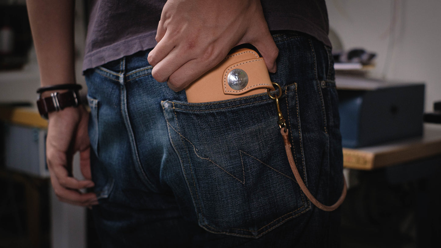 A natural leather wallet with a metal clasp and a leather chain attached. The wallet is in a back pocket of blue jeans.