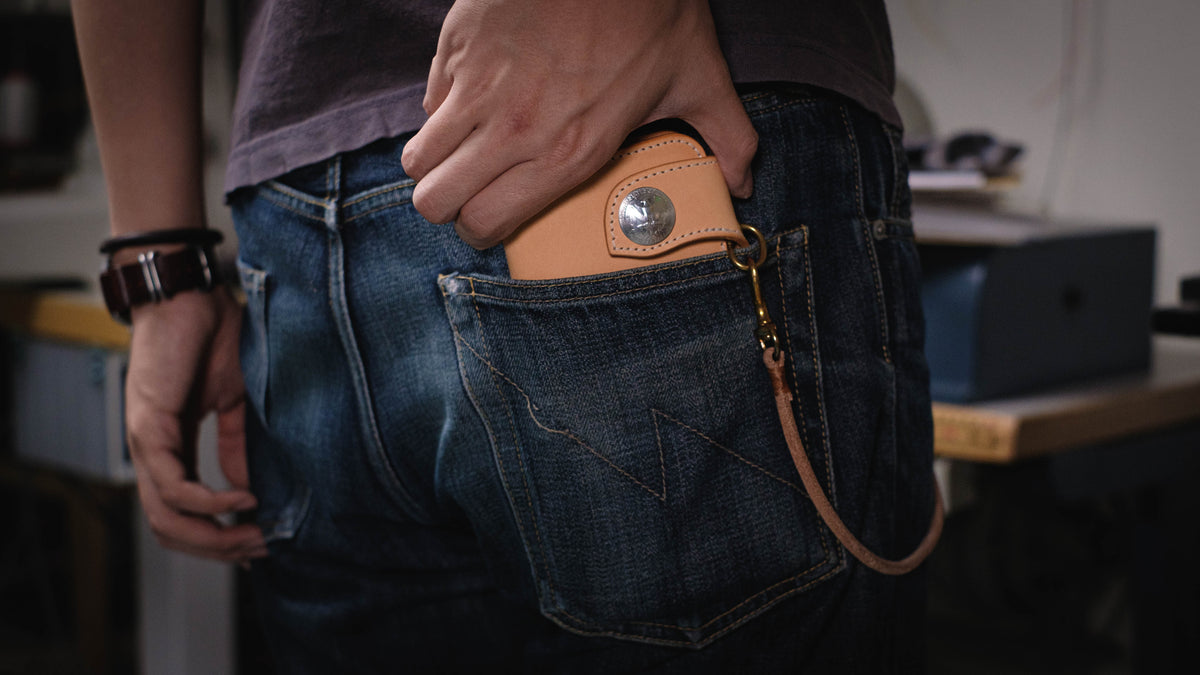 A natural leather wallet with a metal clasp and a leather chain attached. The wallet is in a back pocket of blue jeans.