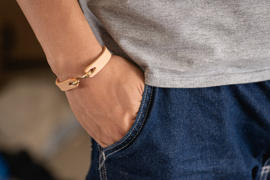 A person wearing a gray t-shirt and blue jeans, with a natural vegetable-tanned leather bracelet featuring a solid brass hook on their wrist.