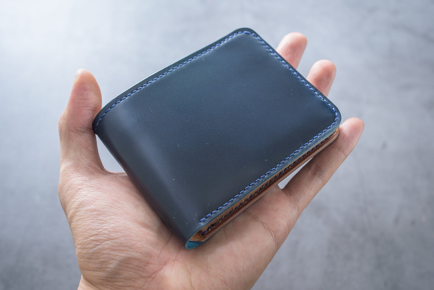 A hand holding a navy blue and whiskey shell cordovan leather billfold wallet.