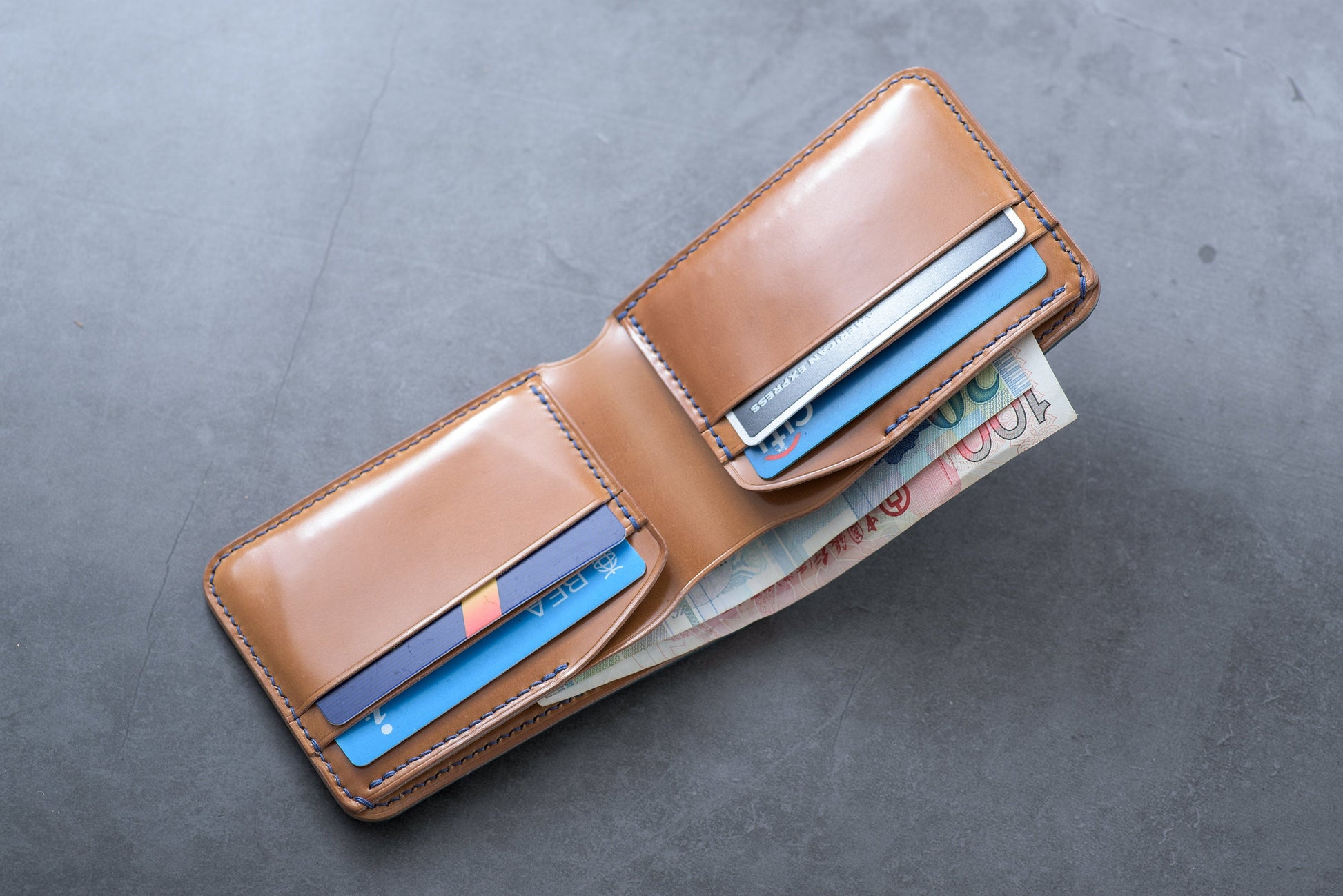 A whisky leather wallet with blue stitching is open, revealing credit cards and cash. The wallet has multiple card slots and a cash compartment.