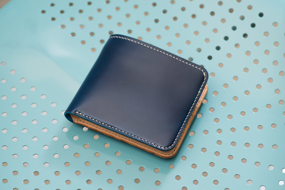 A bi-fold wallet featuring a smooth, dark navy blue leather exterior with contrasting white stitching along the edges. The interior lining and visible side edges are constructed from a natural, light tan-colored leather material.