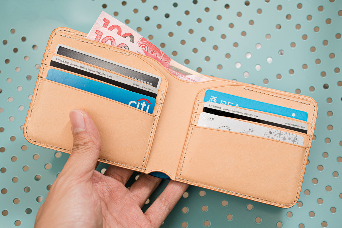 A natural leather bifold wallet featuring multiple interior card slots and a main compartment for currency. The wallet is open, displaying several credit cards and a one hundred dollar banknote tucked inside the billfold section.