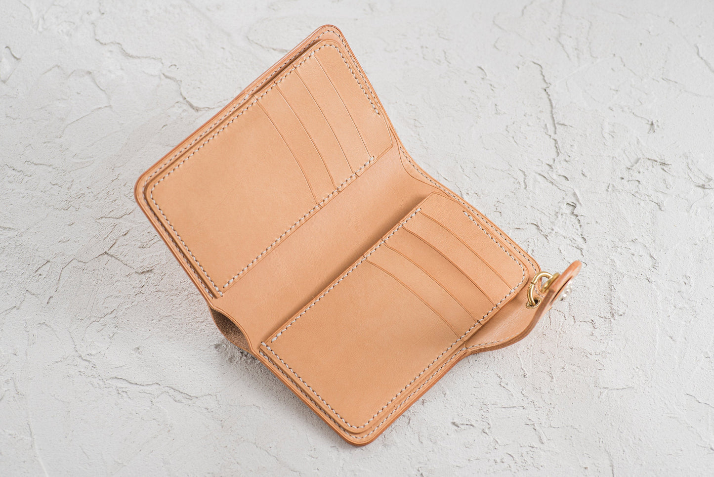 Open, natural leather wallet with 8 card slots and a bill compartment. The wallet is stitched with a contrasting thread and has a gold-colored clasp. The design is minimalist and functional.