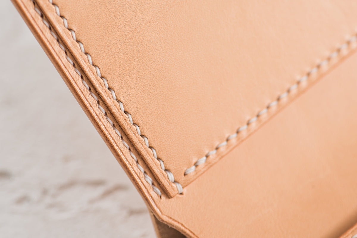 Close-up of a light tan leather item with white stitching. The stitching is visible along the edges, creating a decorative border. The leather appears smooth and the stitching is evenly spaced.