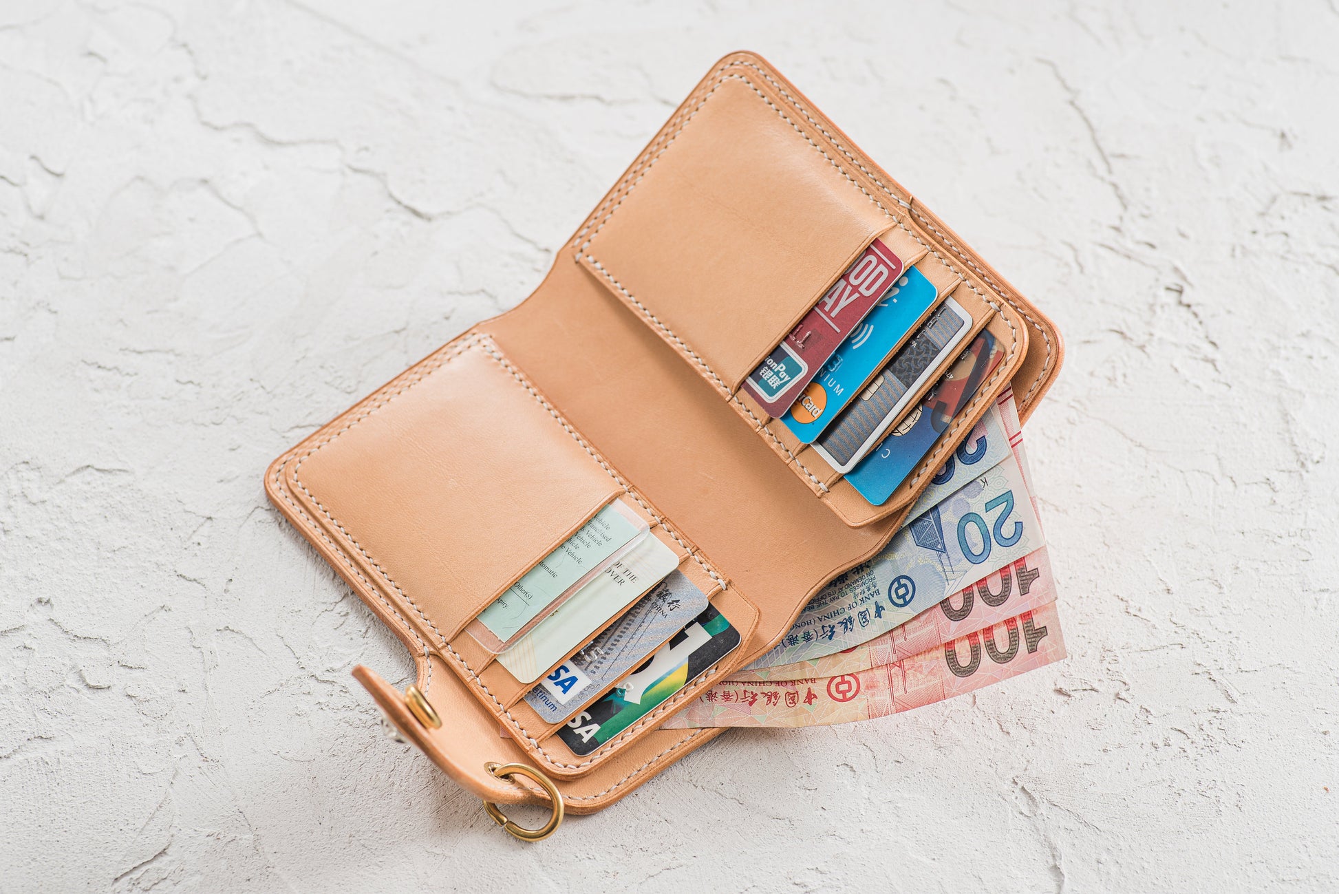 Open, natural leather wallet with 8 card slots and a cash compartment. The wallet is filled with various credit cards and paper currency. A gold-colored key ring is attached to the side.