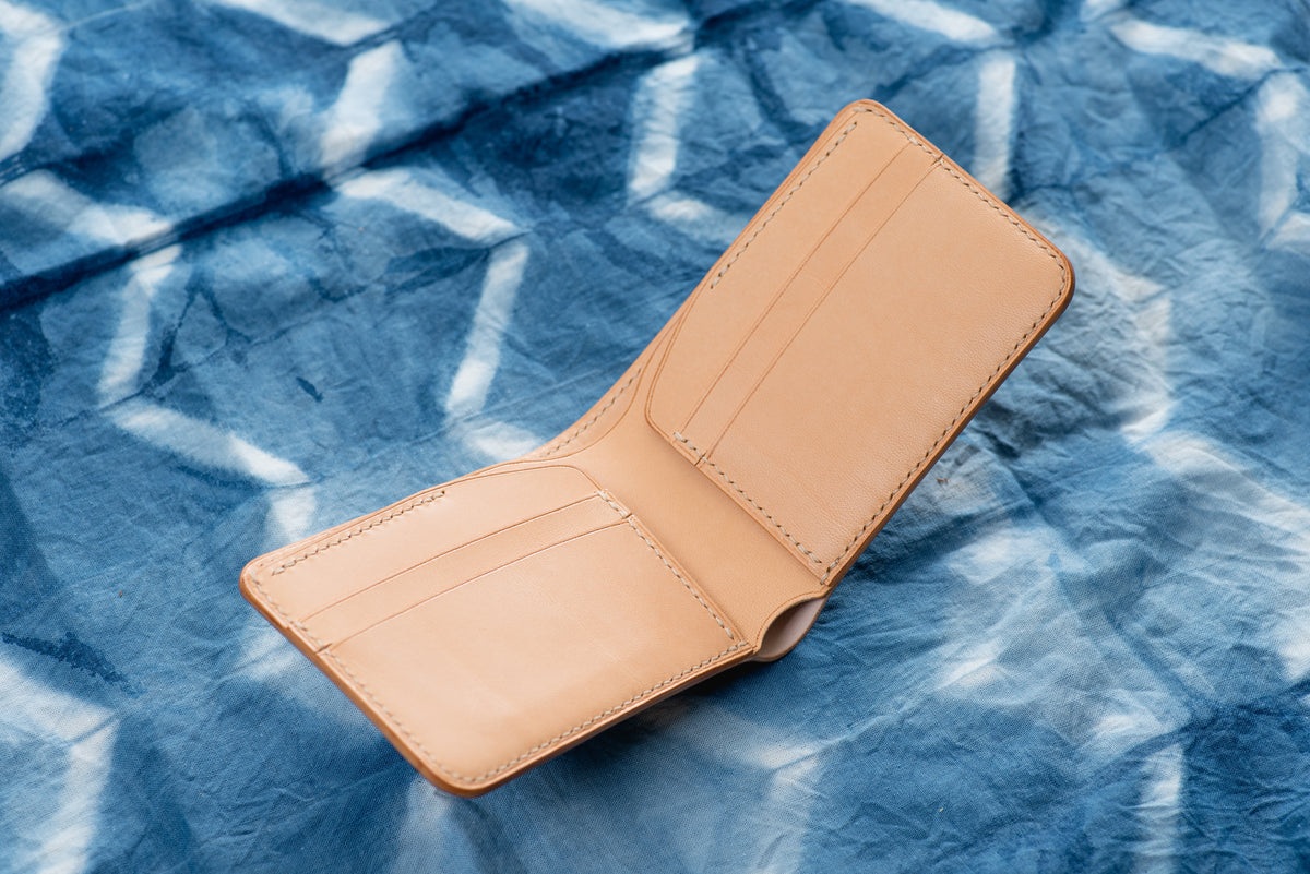 A natural leather bifold wallet featuring visible stitching along the edges, multiple interior card slots, and a main compartment for currency. The wallet is open to display its smooth, natural-colored interior construction.