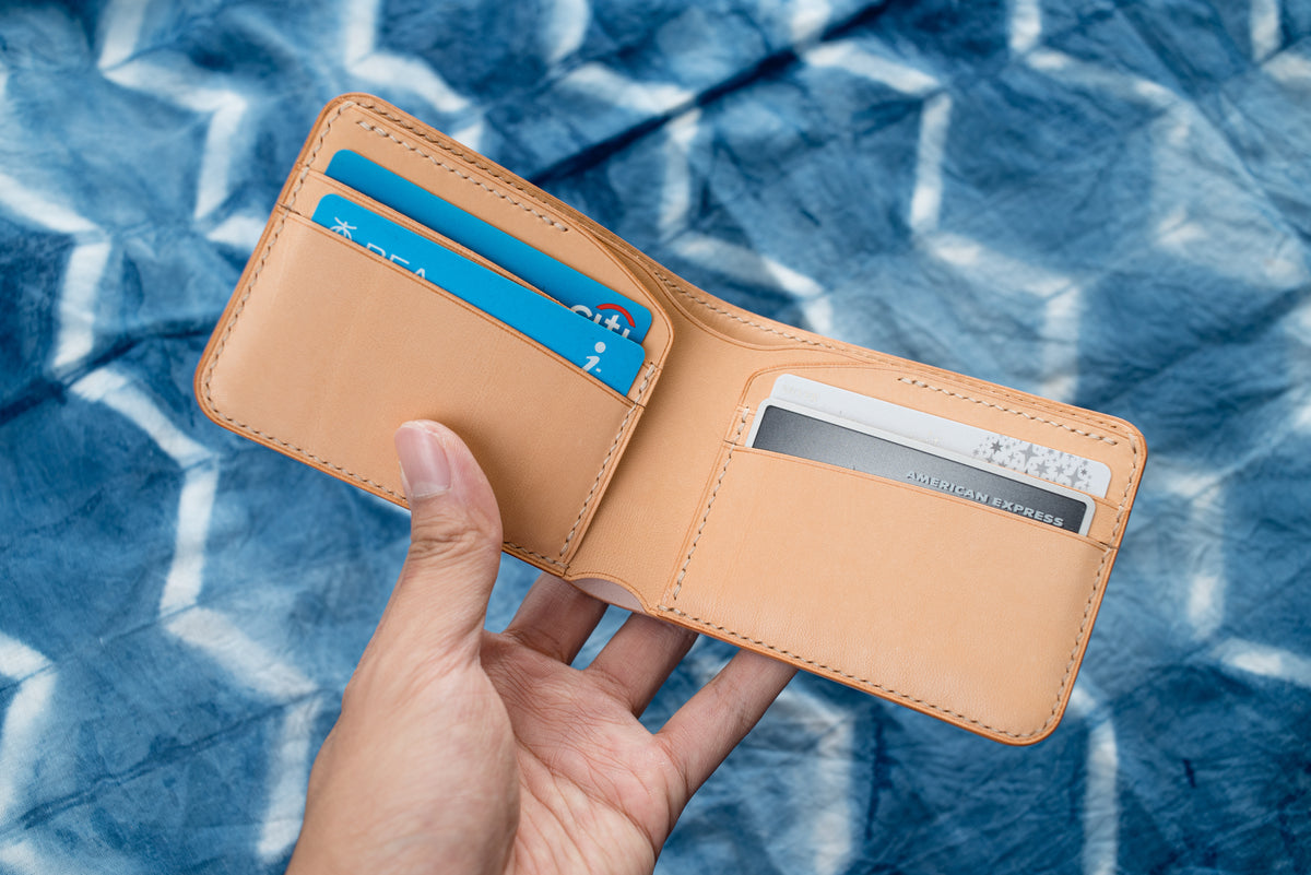 A natural leather bifold wallet featuring visible stitching along the edges, multiple interior card slots, and a large compartment for bills. The wallet contains several credit cards.