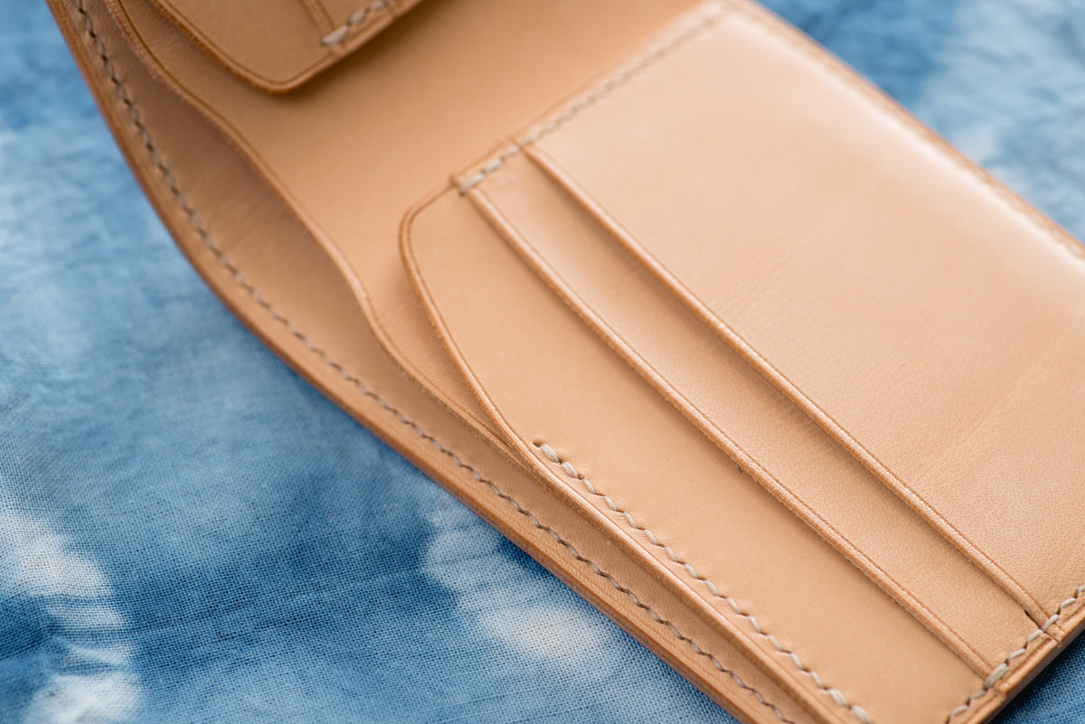 A natural leather bifold wallet featuring visible cream-colored stitching along the edges and interior card slots. The wallet is constructed with smooth, natural-toned leather and includes multiple horizontal slots for organizing cards.